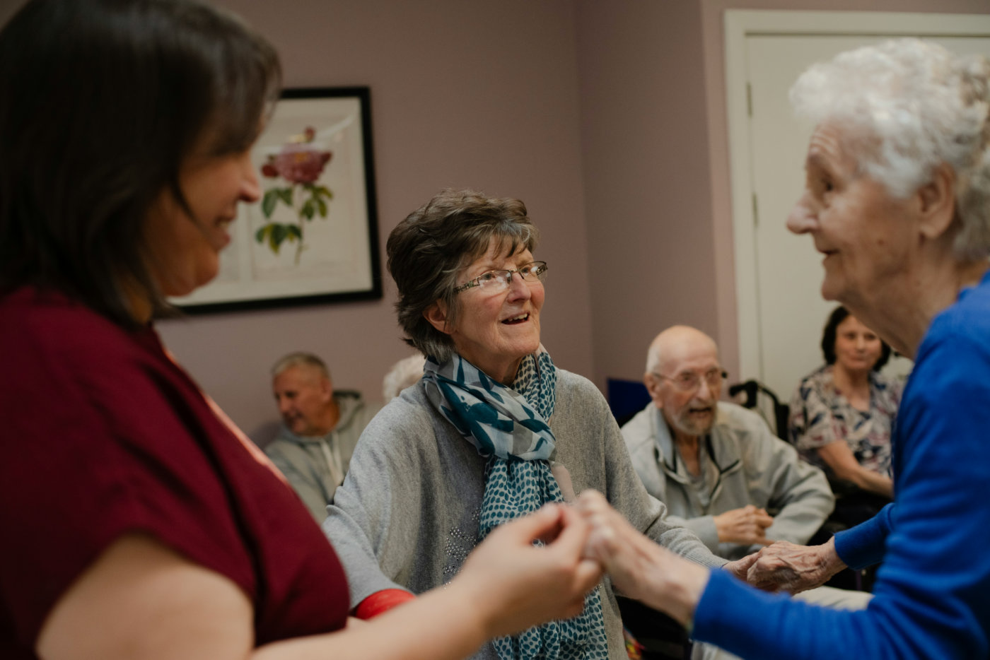 A group of elderly individuals and a caregiver joyfully interact, holding hands, in a cozy room with soft lighting and artwork on the wall.